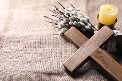 Burning church candle, wooden cross and willow branches on beige cloth, space for text