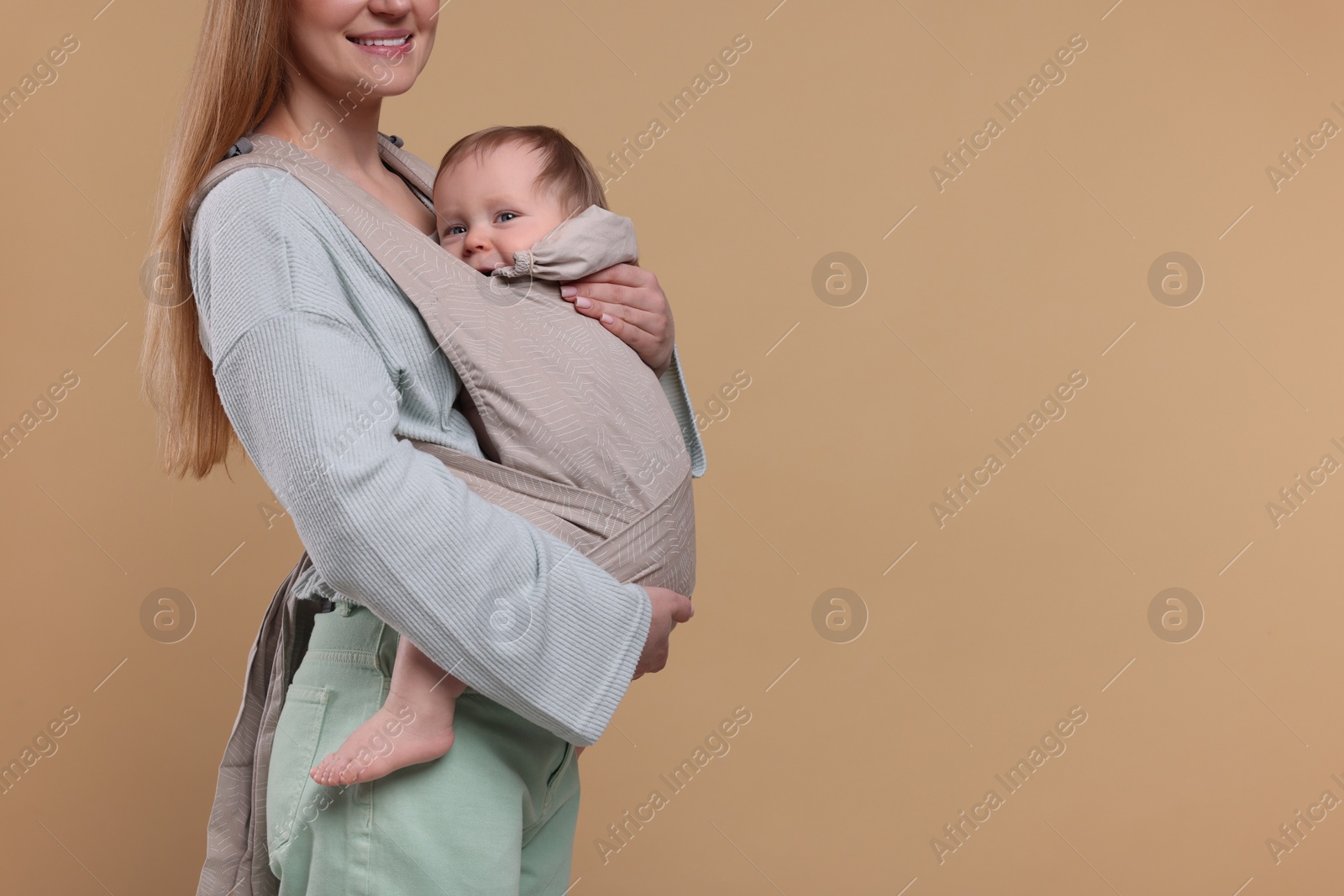 Photo of Mother holding her child in sling (baby carrier) on light brown background, closeup. Space for text