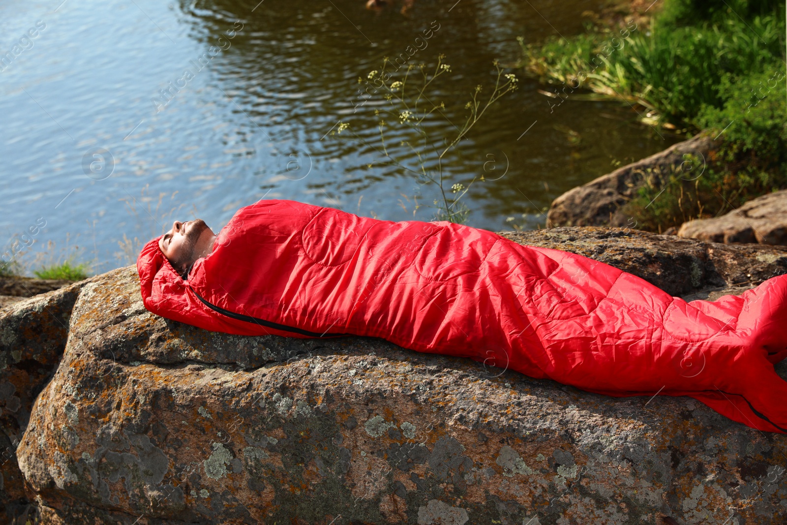 Photo of Young man resting in sleeping bag on cliff near lake