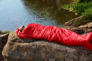 Photo of Young man resting in sleeping bag on cliff near lake