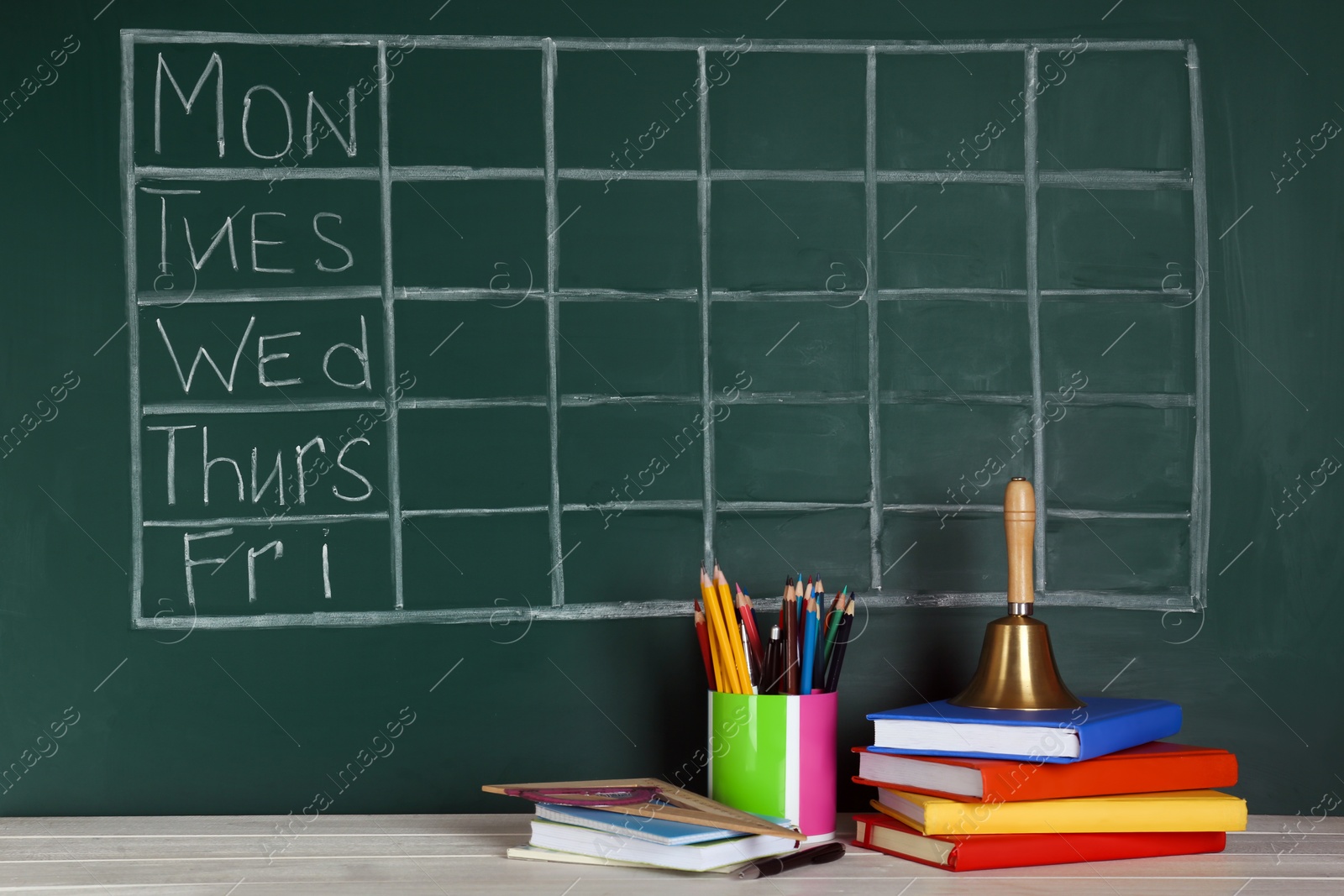 Photo of Bell and different stationery on white wooden table near green chalkboard with drawn school timetable