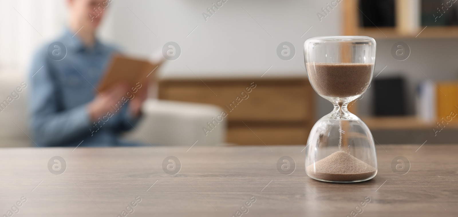 Photo of Hourglass with flowing sand on desk. Man reading book in room, selective focus