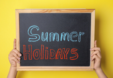 Photo of Schoolgirl holding blackboard with text SUMMER HOLIDAYS on yellow background, closeup