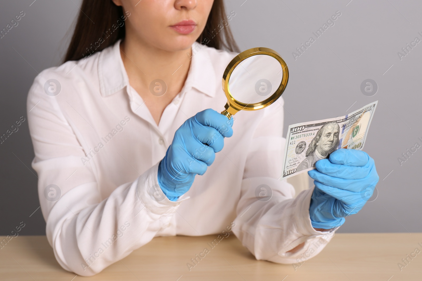 Photo of Expert authenticating 100 dollar banknote with magnifying glass at table on light grey background, closeup. Fake money concept