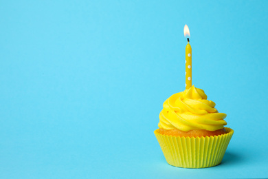 Delicious birthday cupcake with yellow cream and burning candle on light blue background. Space for text