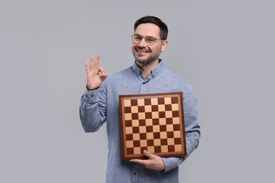 Smiling man holding chessboard and showing OK gesture on light grey background