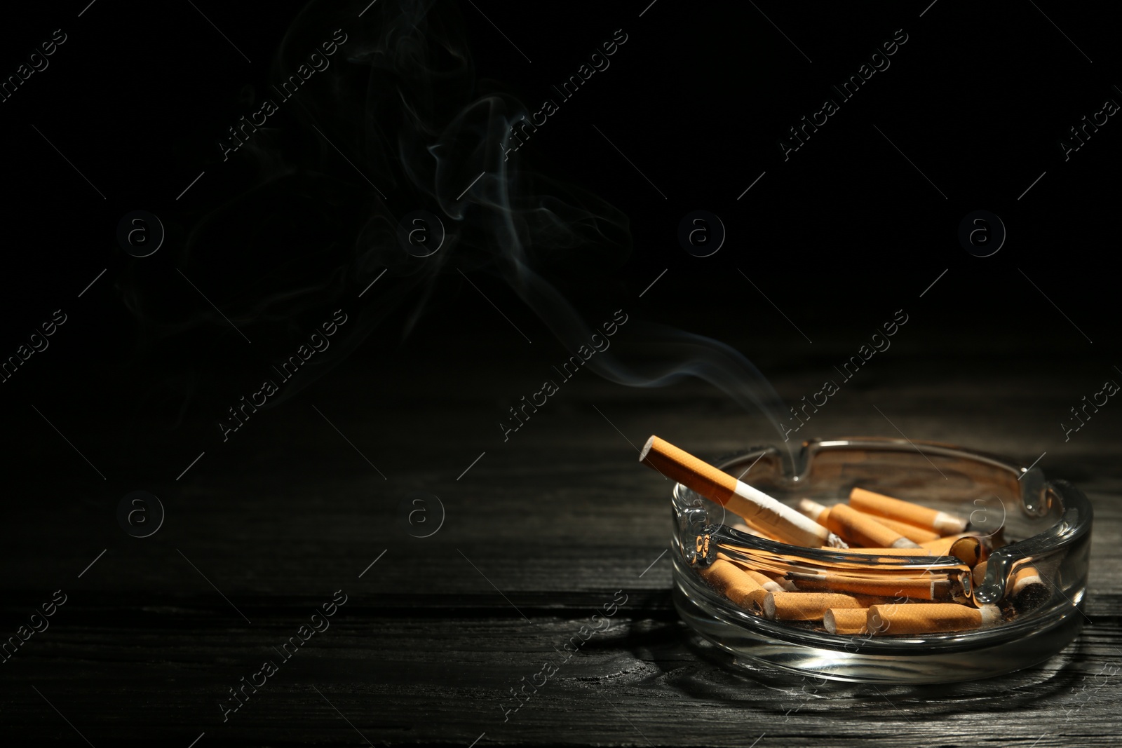 Photo of Smoldering cigarette in glass ashtray on dark wooden table against black background. Space for text