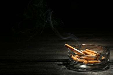 Smoldering cigarette in glass ashtray on dark wooden table against black background. Space for text