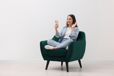 Photo of Beautiful woman having video chat via smartphone while sitting in armchair near white wall indoors