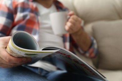 Photo of Man with cup of drink reading magazine on sofa, closeup
