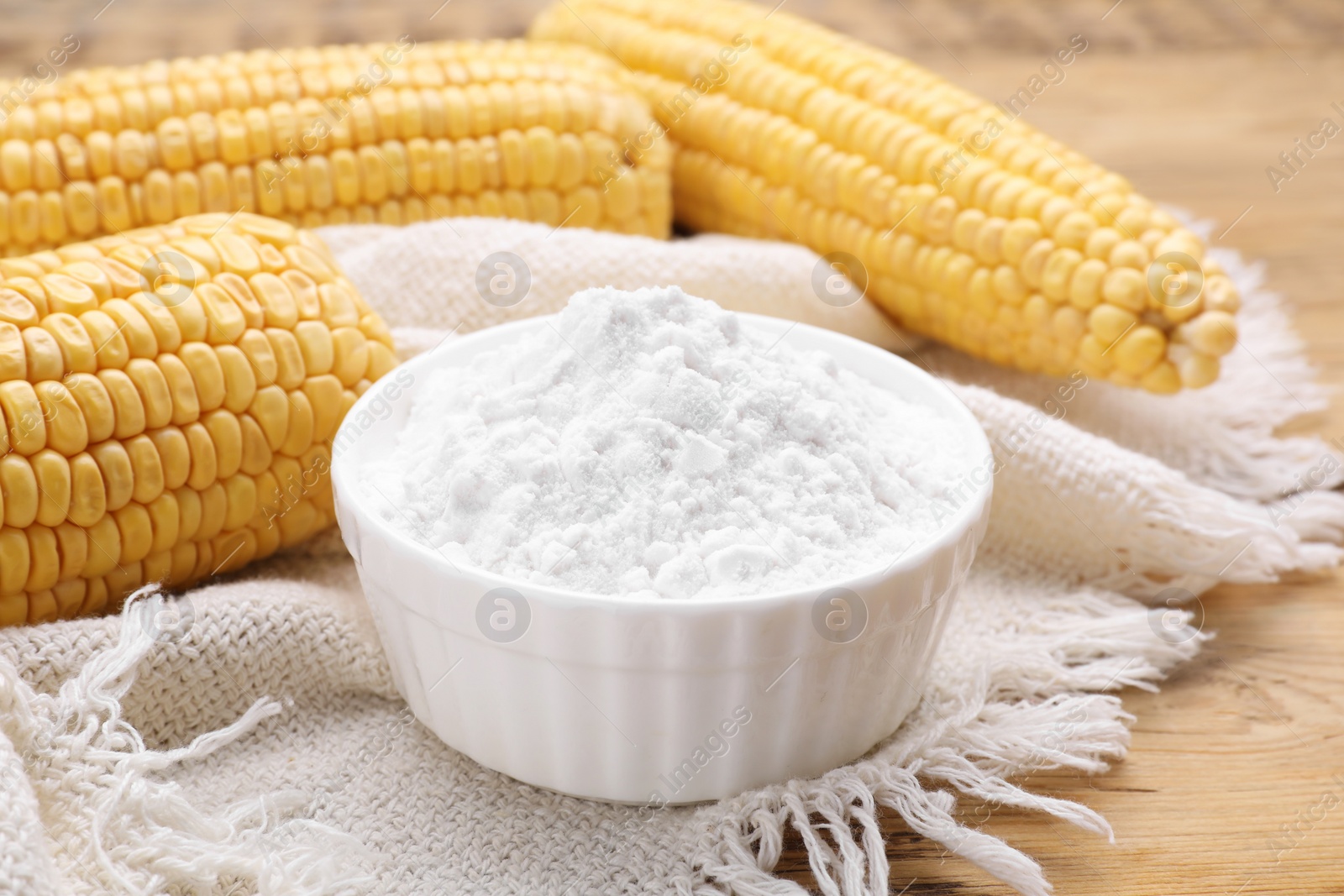 Photo of Bowl with corn starch and ripe cobs on wooden table, closeup