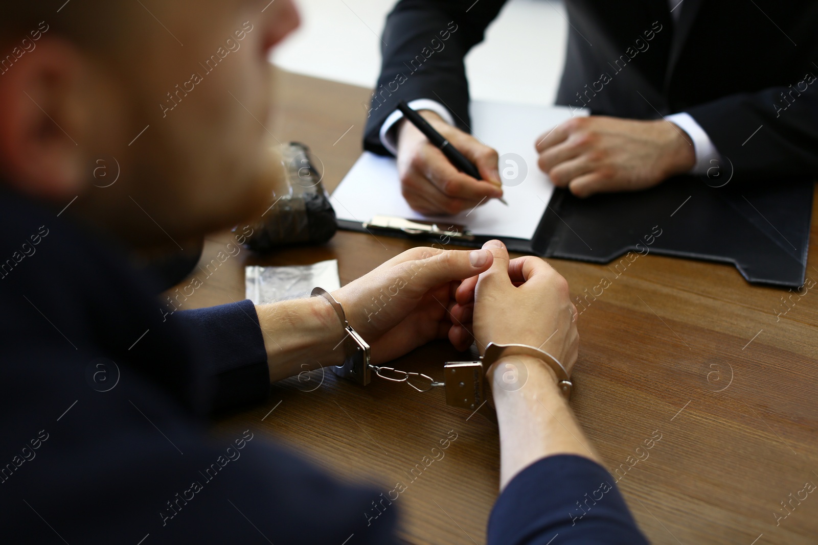 Photo of Police officer interrogating criminal in handcuffs at desk indoors