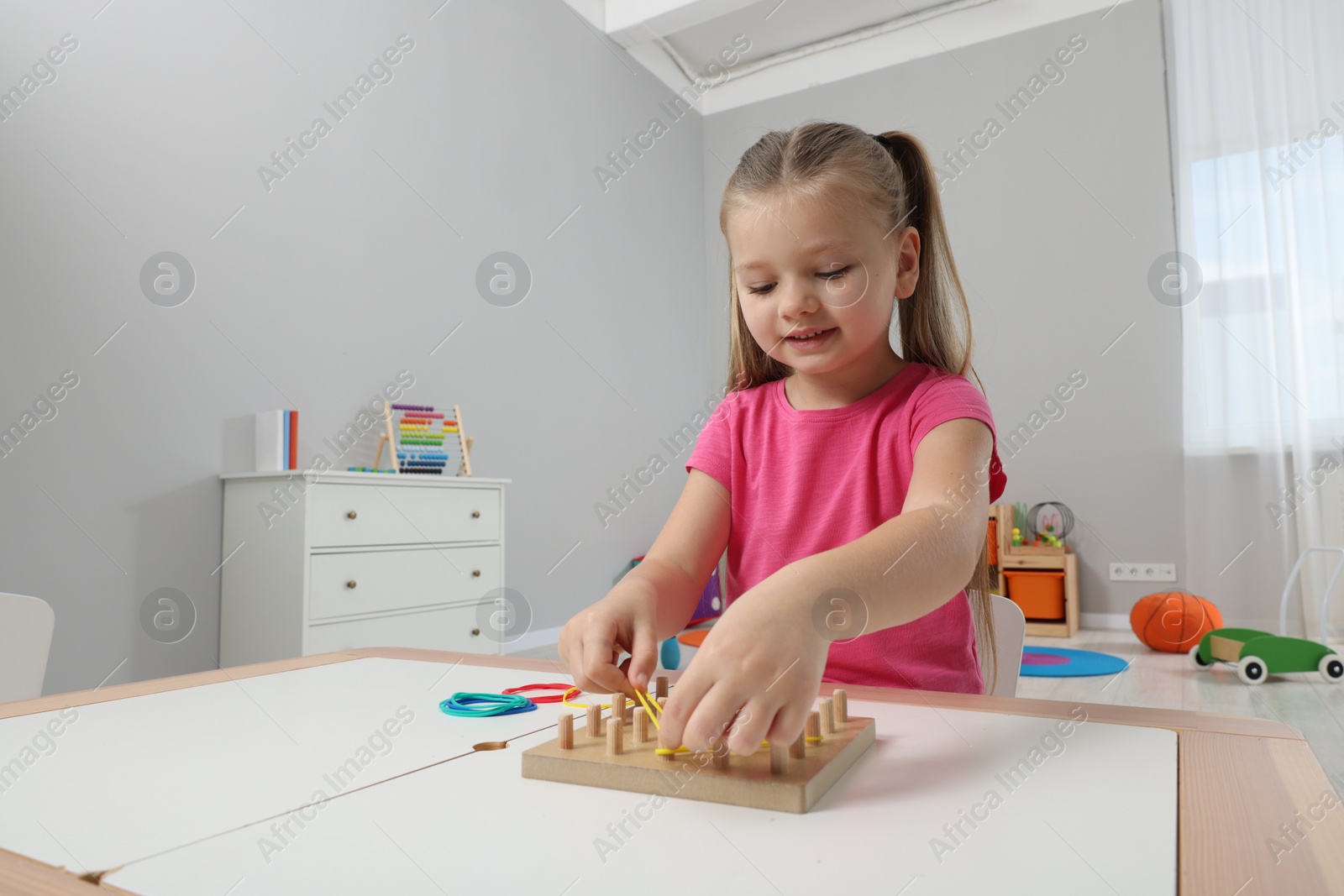Photo of Motor skills development. Girl playing with geoboard and rubber bands at white table in kindergarten. Space for text