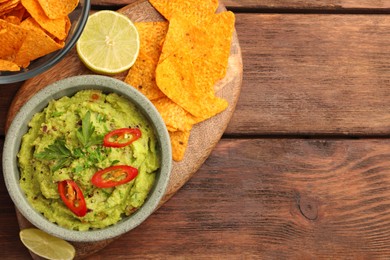 Bowl of delicious guacamole, lime and nachos chips on wooden table, top view. Space for text