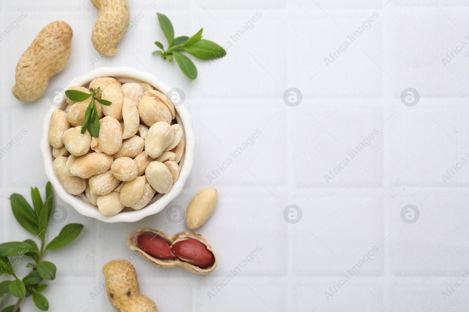 Photo of Fresh peeled peanuts in bowl and leaves on white tiled table, flat lay. Space for text