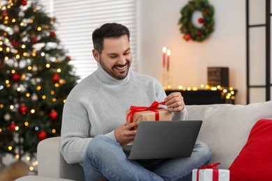 Photo of Celebrating Christmas online with exchanged by mail presents. Smiling man opening gift box during video call on laptop at home