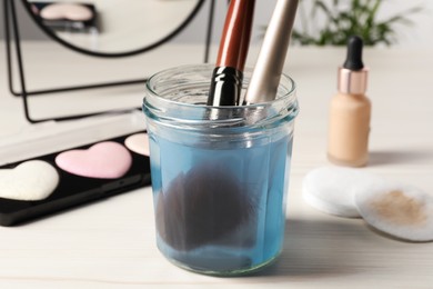 Photo of Cleaning makeup brushes in jar with special liquid on white wooden table, closeup