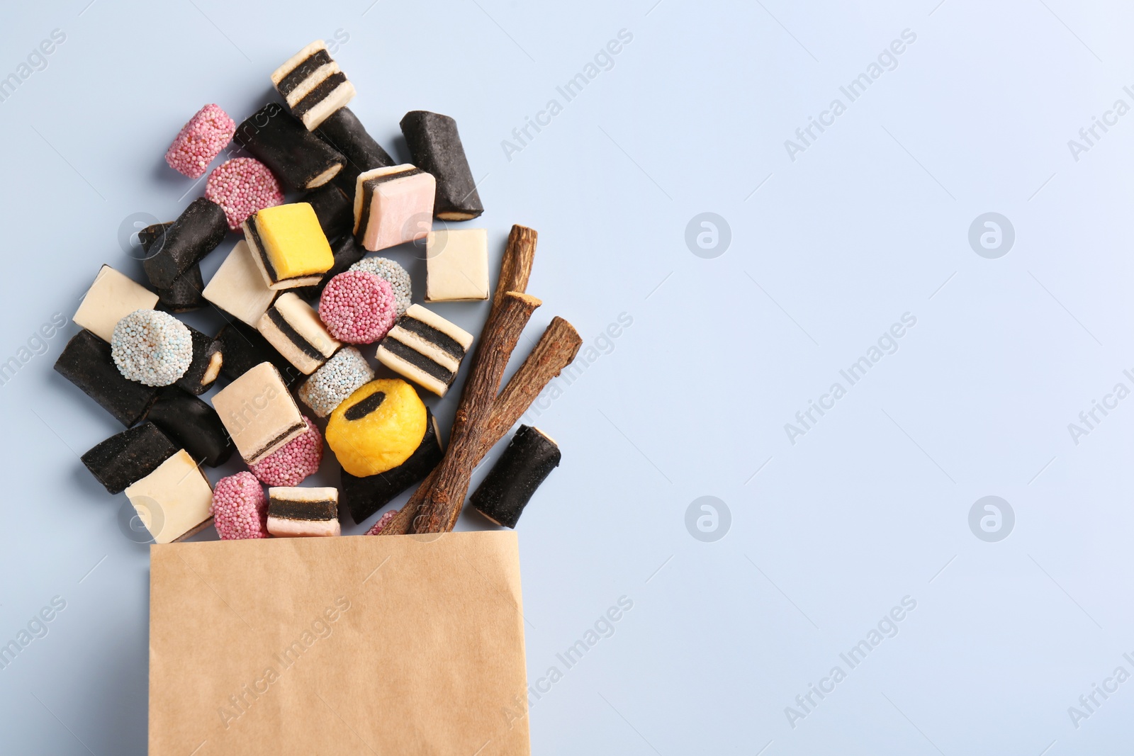 Photo of Paper bag with tasty candies and dried cut liquorice root on light blue background, top view. Space for text