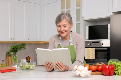 Photo of Happy woman with recipe book at table in kitchen