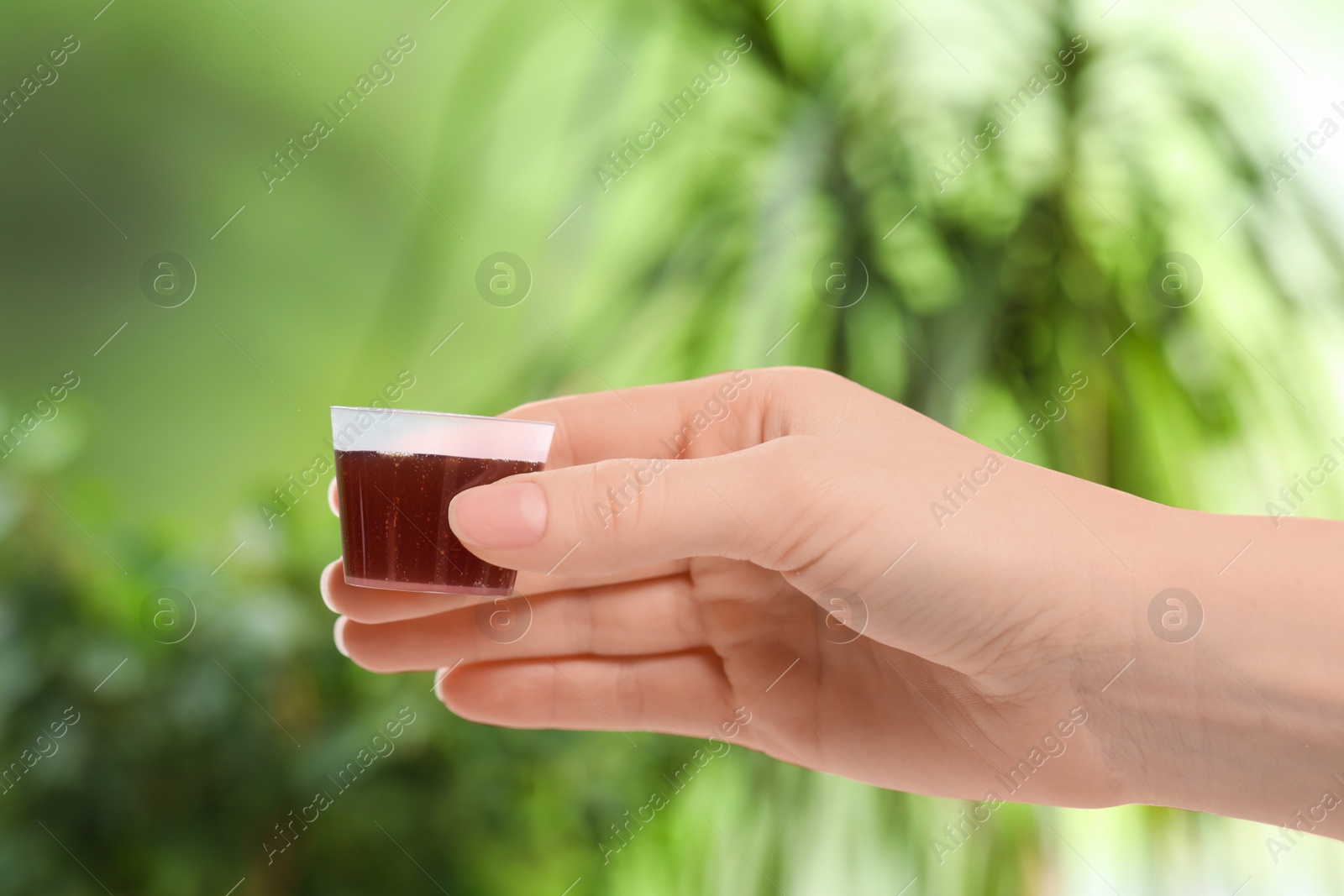 Photo of Woman holding measuring cup with syrup on blurred background, closeup. Cold medicine