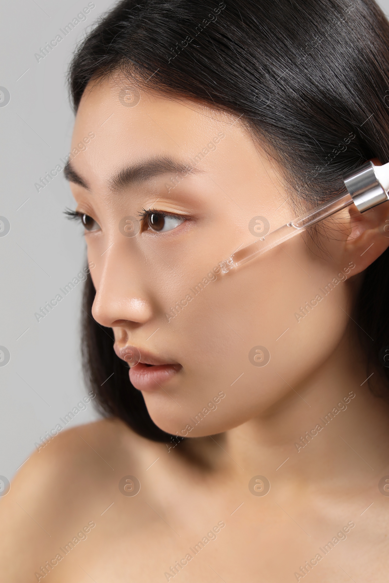 Photo of Beautiful young woman applying cosmetic serum onto her face on grey background, closeup