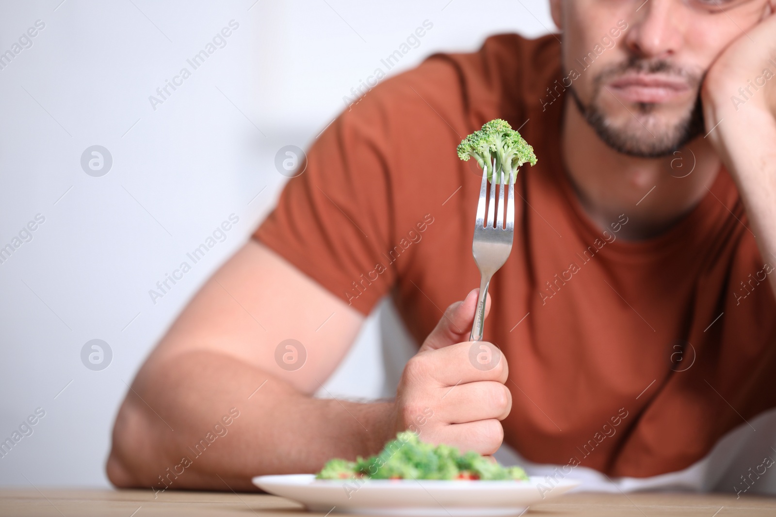 Photo of Unhappy man with broccoli on fork at table, closeup