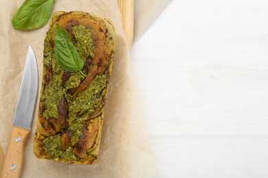 Freshly baked pesto bread with basil and knife on white wooden table, flat lay. Space for text