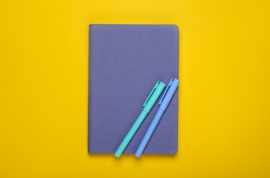 Photo of Closed blue notebook and pens on yellow background, top view