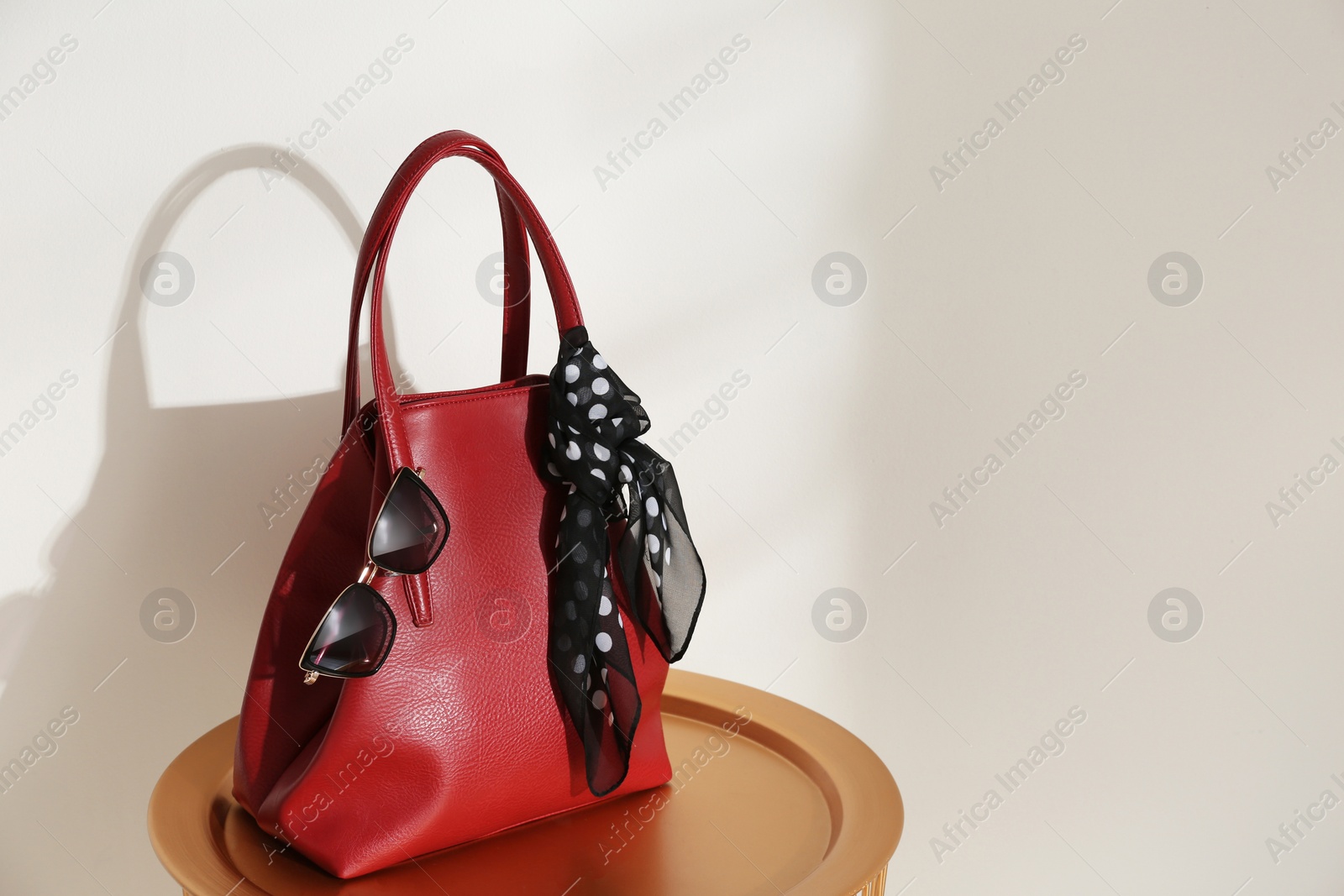 Photo of Stylish red woman's bag, scarf and sunglasses on table near light wall. Space for text