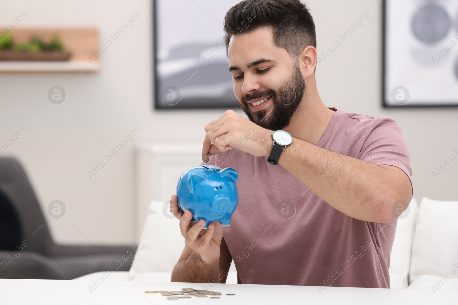 Photo of Happy young man putting money into piggy bank at white table indoors