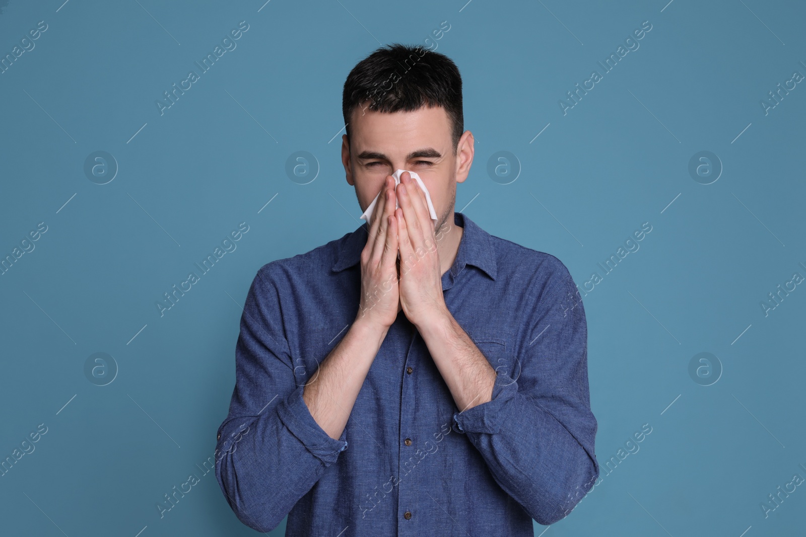 Photo of Young man blowing nose in tissue on blue background. Cold symptoms