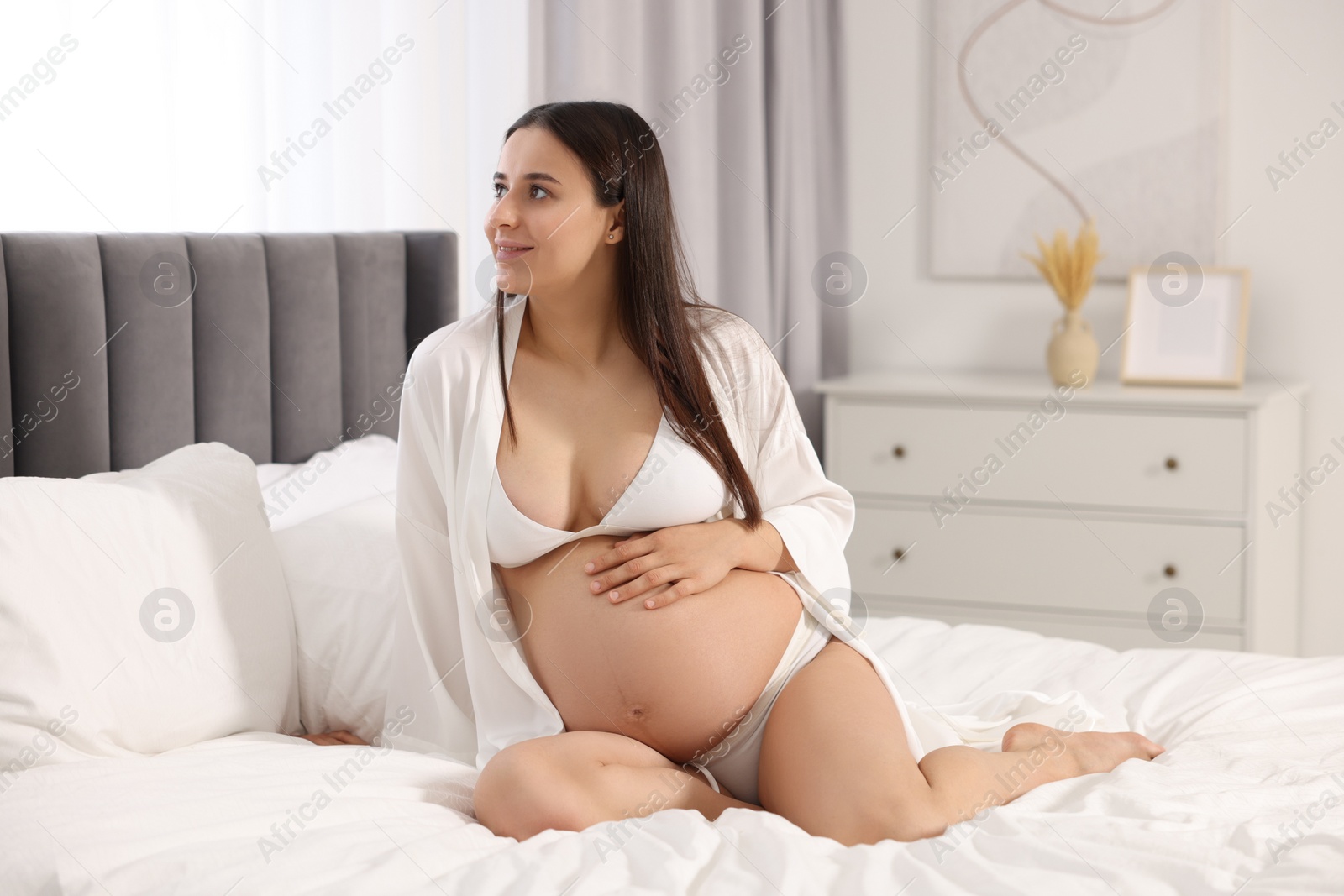 Photo of Beautiful pregnant woman in stylish comfortable underwear and robe on bed at home