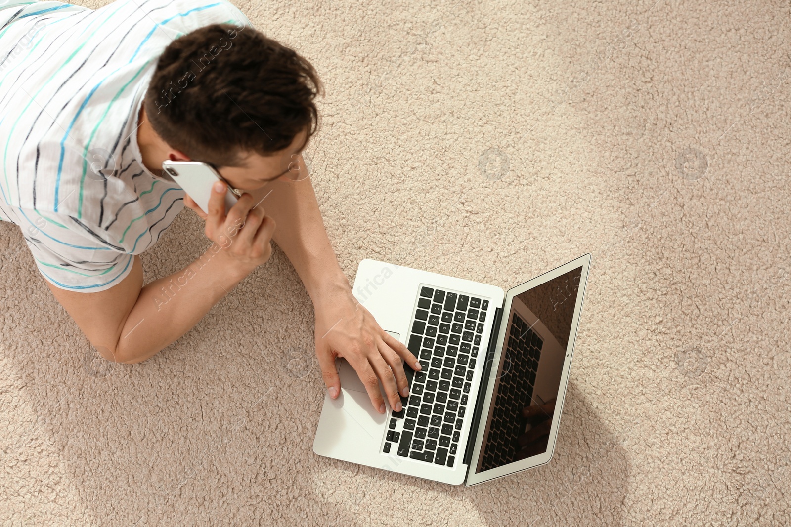 Photo of Handsome young man with mobile phone and laptop on cozy carpet at home