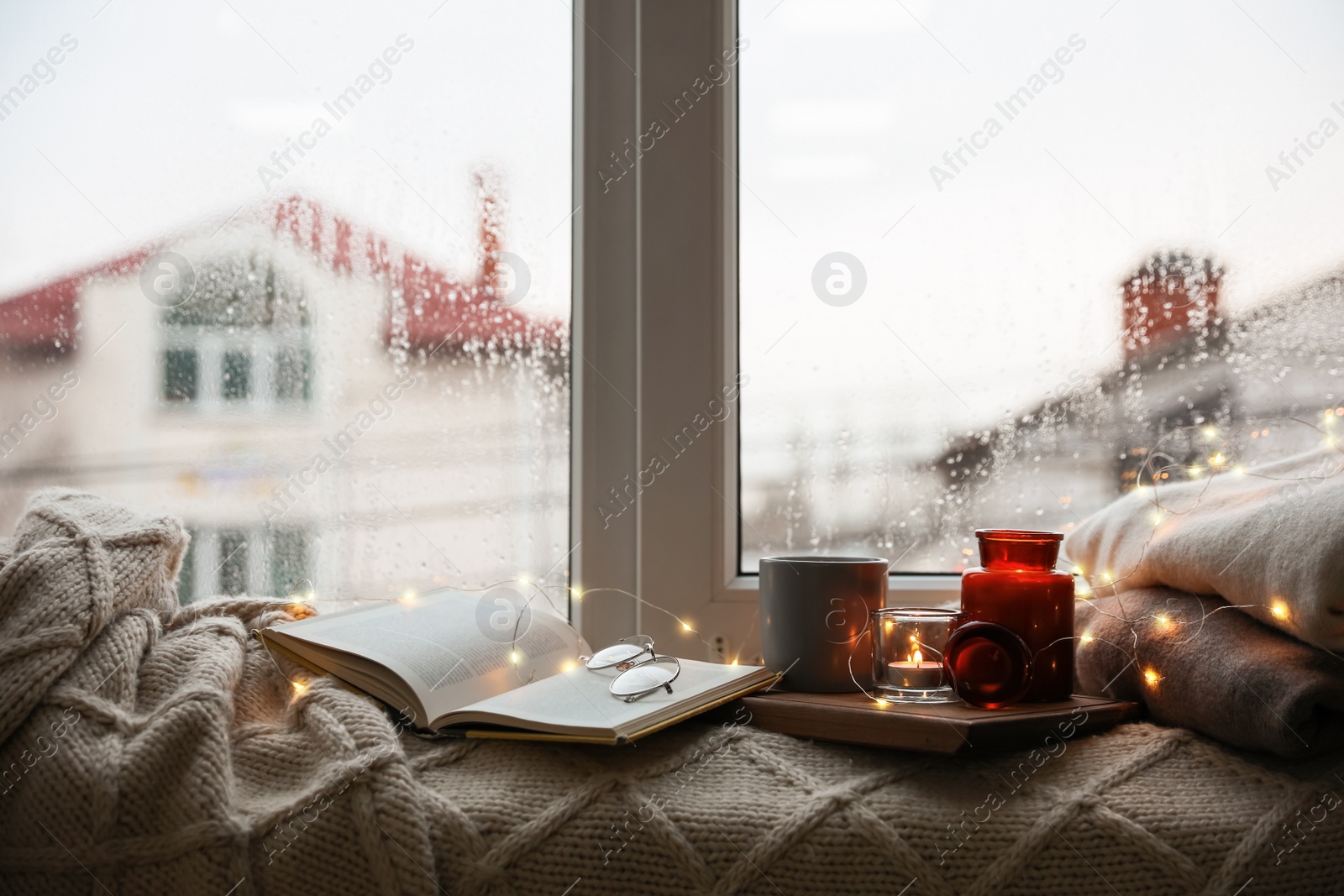 Photo of Cup of hot drink, open book and Christmas lights on knitted blanket near window. Cozy season