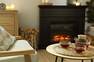 Teapot and cups of drink on coffee table near stylish fireplace in cosy living room. Interior design