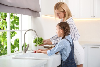 Photo of Little girl with her mother washing vegetables together in modern kitchen