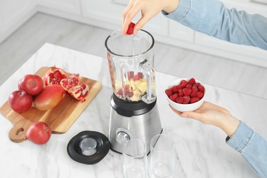 Photo of Woman adding raspberry into blender with ingredients for smoothie at white table, closeup