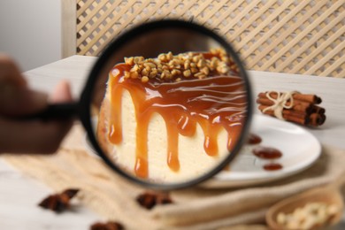 Woman looking through magnifying glass at caramel cheesecake on white table, closeup