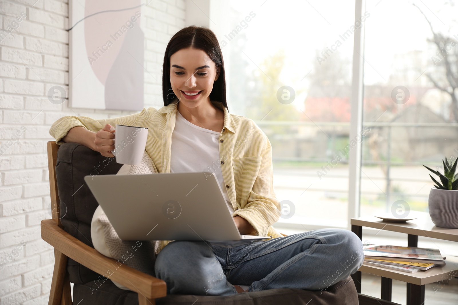 Photo of Beautiful young woman with cup of drink using laptop at home