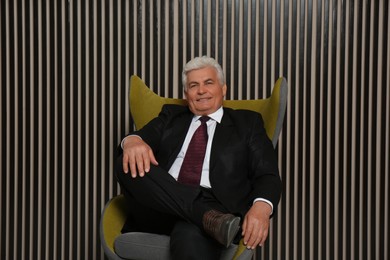 Happy mature businessman sitting in armchair near wood slat wall