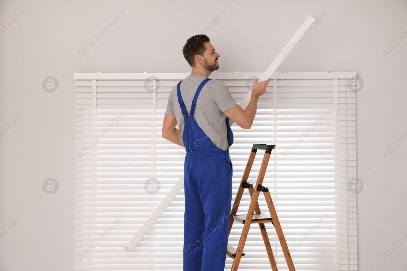 Photo of Worker in uniform installing horizontal window blinds on stepladder indoors