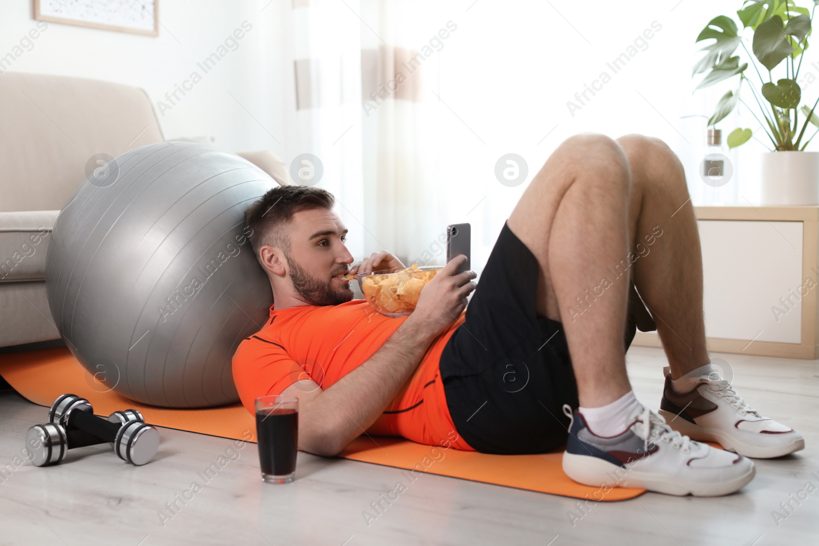 Photo of Lazy young man with smartphone eating junk food on yoga mat at home
