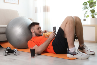Photo of Lazy young man with smartphone eating junk food on yoga mat at home