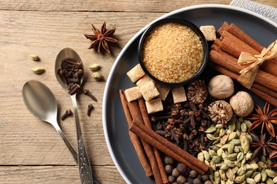 Plate with different aromatic spices and spoons on wooden table, flat lay