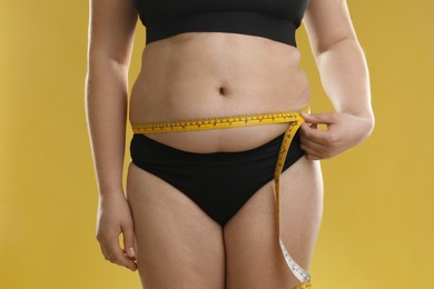 Woman measuring belly with tape on goldenrod background, closeup. Overweight problem