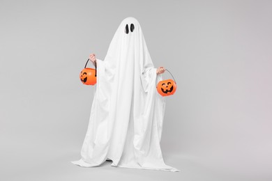 Child in white ghost costume holding pumpkin buckets on light grey background. Halloween celebration