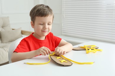 Little boy tying shoe lace using training cardboard template at white table indoors