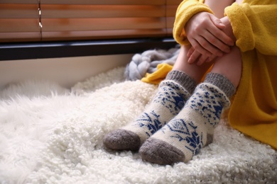 Photo of Woman wearing knitted socks on window sill indoors, closeup. Warm clothes