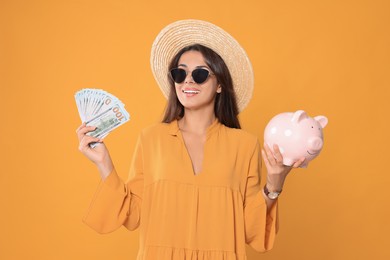 Young woman in stylish sunglasses, straw hat with money and piggy bank on orange background