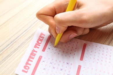 Photo of Woman filling out lottery ticket with pen on wooden table, closeup. Space for text